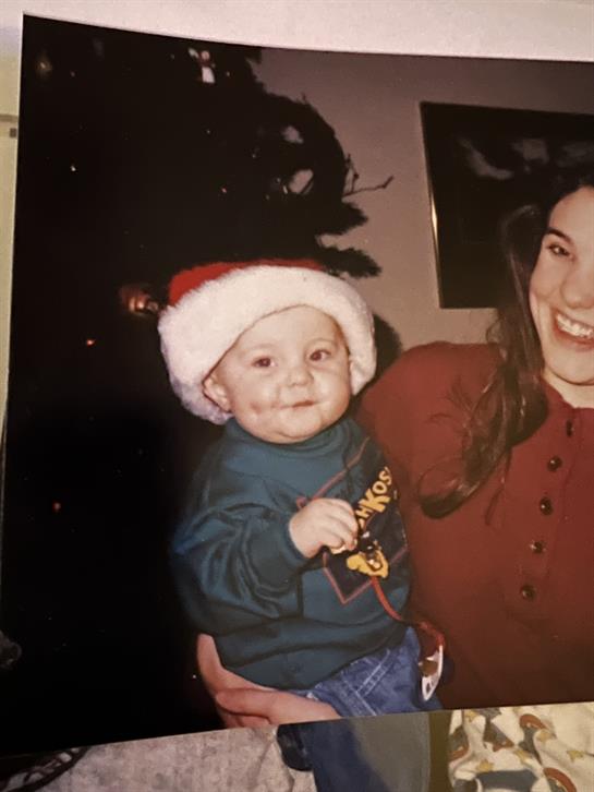A joyful baby wearing a Santa hat is held by his smiling mother during a festive home gathering.