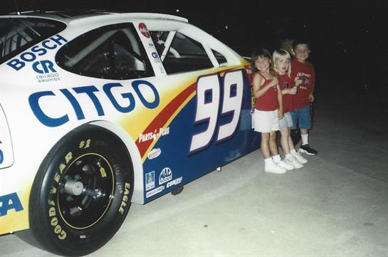Kids stand beside a colorful NASCAR race car, excitedly posing during a community event.