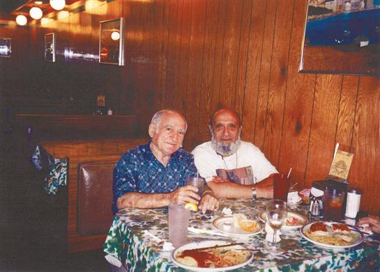 Two men sit at a table, surrounded by plates of food and drinks, enjoying their time together.