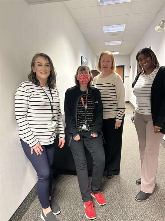 Four women stand together, smiling, in a professional hallway, enjoying a casual moment at work.