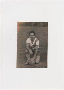 A young man kneels in a grassy field, holding a basketball and gazing at the camera.