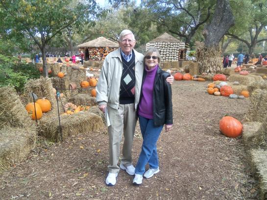 An elderly couple poses happily among pumpkins and hay bales at an autumn festival.