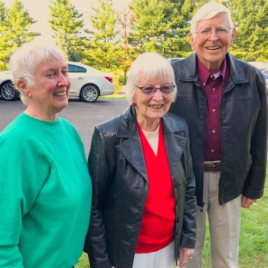 Three older adults smile and engage in conversation outdoors near greenery and parked cars.