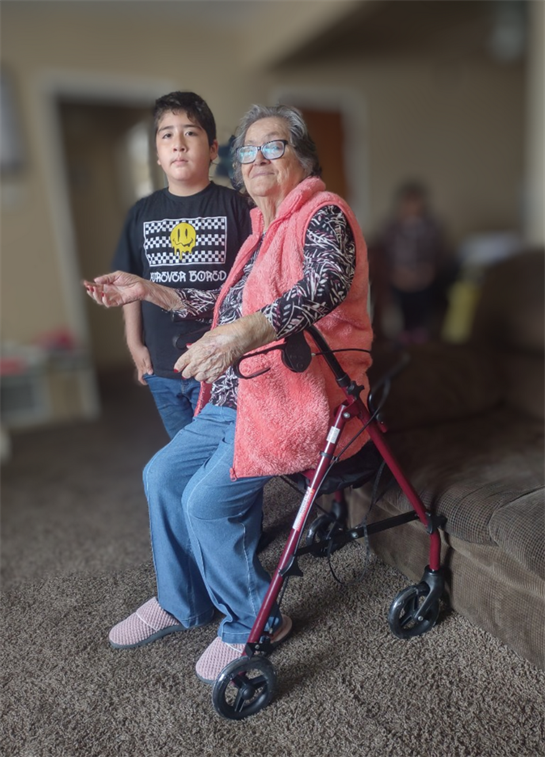 An elderly woman sits on a couch with a walker, smiling at a young boy beside her in a cozy room.