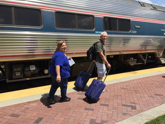 Two travelers pull their luggage while walking towards a train during a bright day.