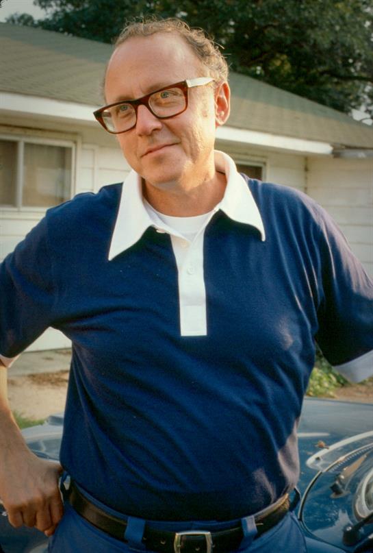 Man with glasses stands confidently by a car outside a house, exuding a casual and relaxed vibe.