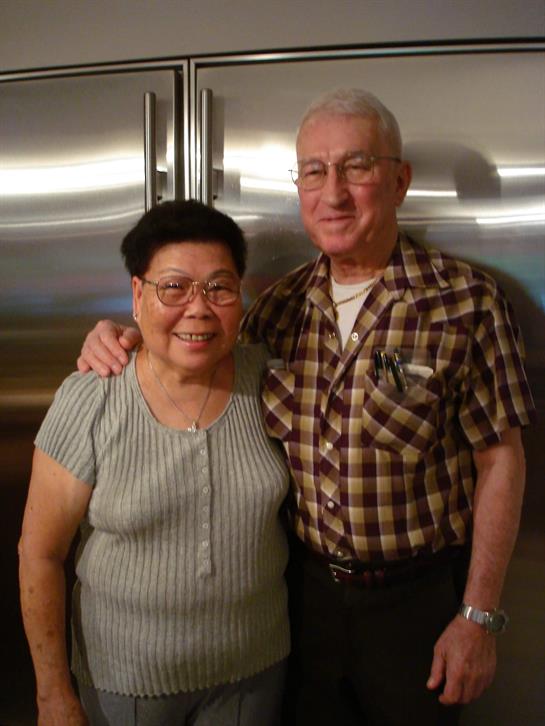 A happy elderly couple stands together in their kitchen, enjoying family time.