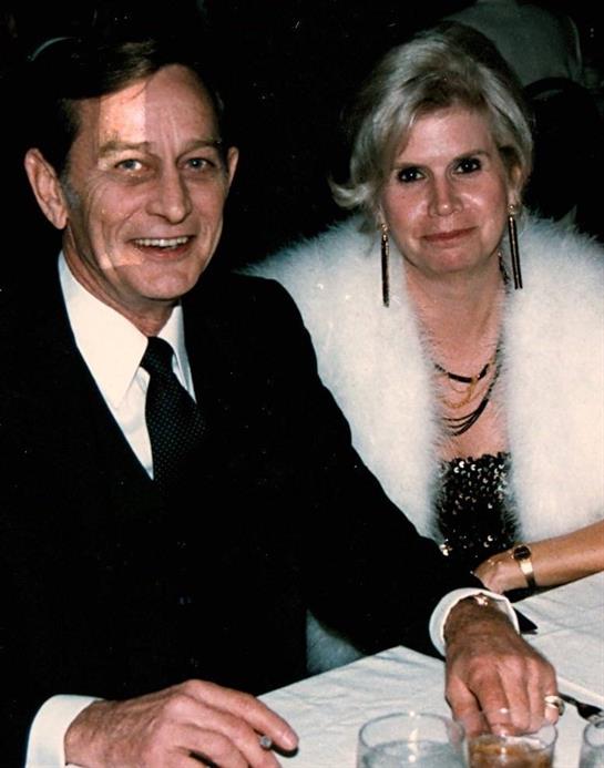 A couple dressed elegantly smiles while seated at a table during an evening event.