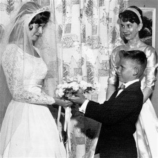 A bride in a classic dress is presented with flowers by a young boy, while a woman stands nearby.