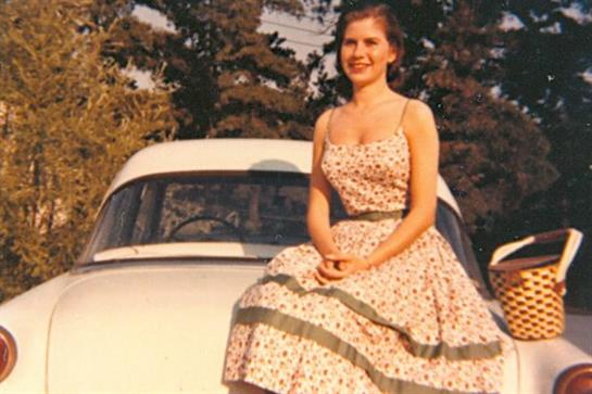 A young woman in a floral dress poses gracefully on a vintage car, surrounded by trees and sunlight.