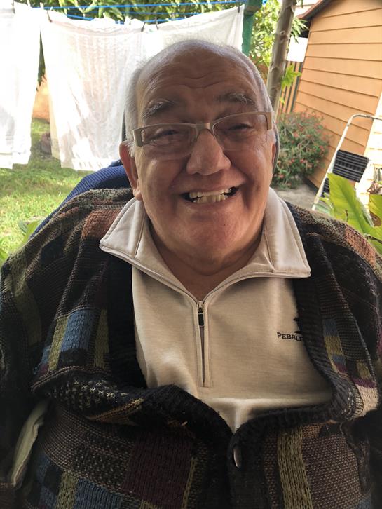 An elderly man sits in a garden, smiling brightly while wearing a comfortable sweater.