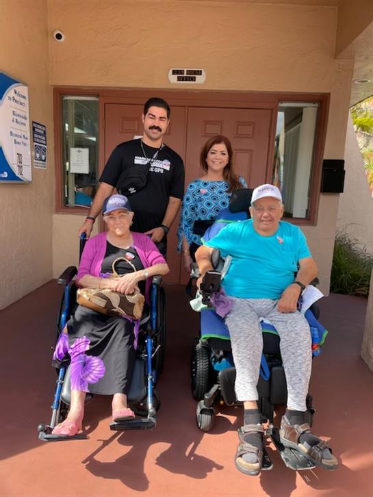 Seniors in wheelchairs smile as volunteers assist them outside a community center.