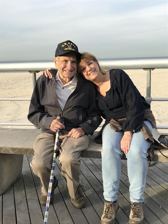 Two individuals sit together on a boardwalk, sharing a warm moment by the beach.