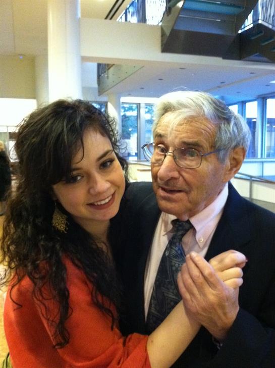 A young woman smiles warmly while holding hands with an older man at a lively event.
