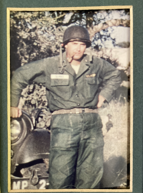 A soldier stands proud with hands on hips near a military vehicle surrounded by nature.