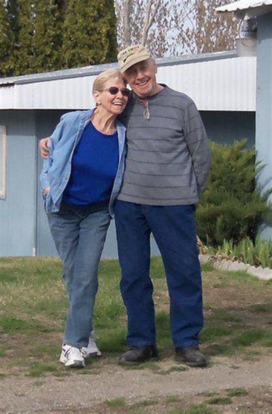 A couple stands close outside their home, smiling in casual, comfortable clothes.