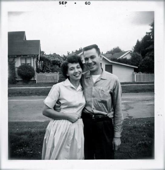 A couple stands close together, smiling warmly in a suburban neighborhood on a bright day.