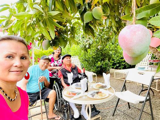 Group of friends gather for a joyful meal under a mango tree in a garden on a sunny day.