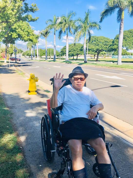 A man waves cheerfully while seated in a wheelchair on a sunny urban street.
