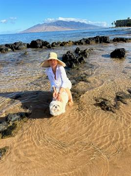 A woman wearing a sun hat enjoys time with her small dog in shallow coastal water while exploring.