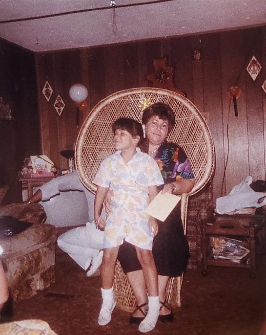 Child in a light outfit sits on a chair with a smiling caregiver in a bright blouse.