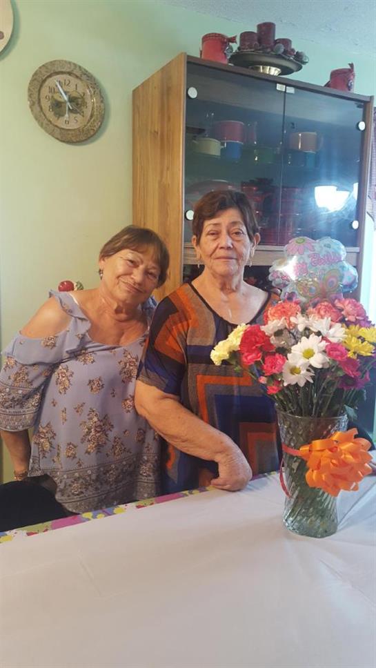 Two women with warm smiles stand side by side next to a vibrant bouquet, enjoying a friendly moment.