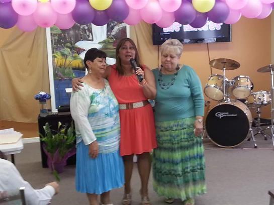 Three women enjoy a lively celebration, singing together in a decorated hall filled with balloons.