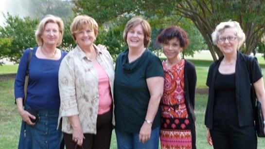 Four women chat and smile together in a sunny, grassy park surrounded by trees.