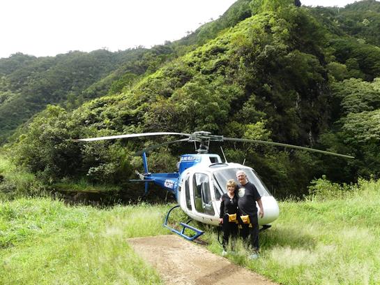 Two people stand by a helicopter in a lush, green landscape, ready for their journey.