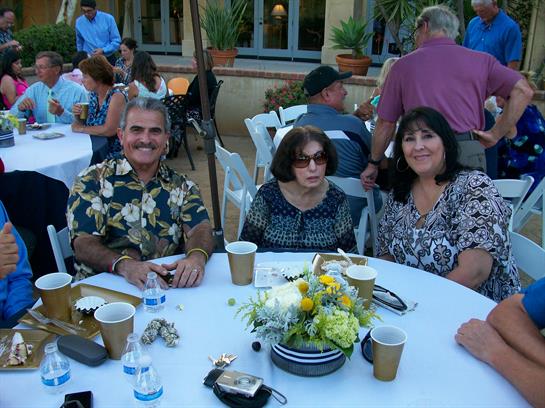 A group of friends sits at a round table sharing smiles and drinks during a lively outdoor event.