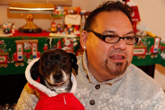 A man holds a small dog in a red outfit while celebrating the holiday season indoors.