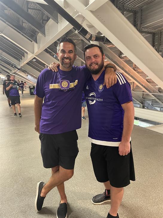 Two friends pose together wearing purple jerseys, smiling in a lively stadium environment.