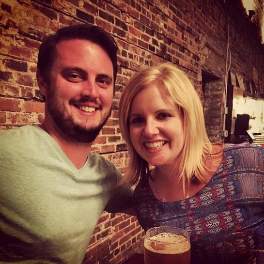 Couple poses for a cheerful photo while sharing drinks at a brick-walled bar during the night.