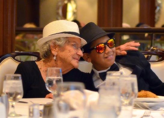 Two cheerful seniors sit together at a banquet table, both dressed in stylish hats and sunglasses.