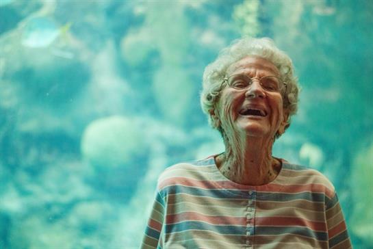 A joyful elderly woman stands smiling amid a colorful turquoise backdrop, enjoying her moment.