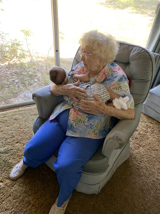 An elderly woman holds her newborn grandchild with warmth in a sunny living room.