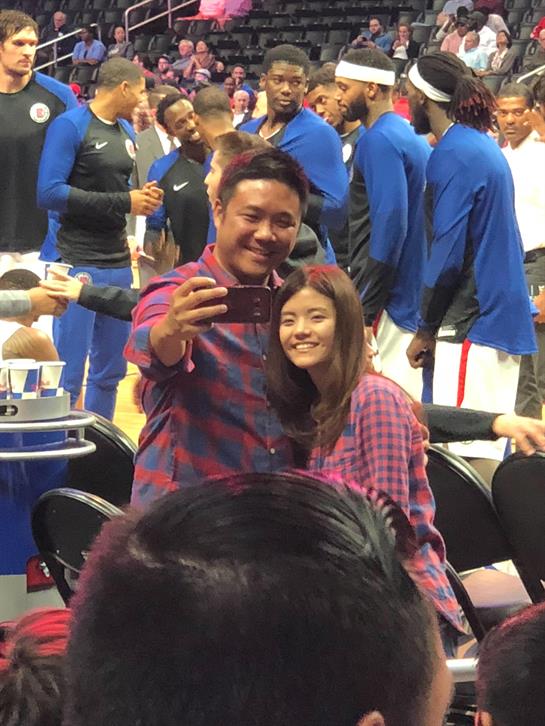 A man and a woman smile together for a selfie at a lively basketball game with players nearby.