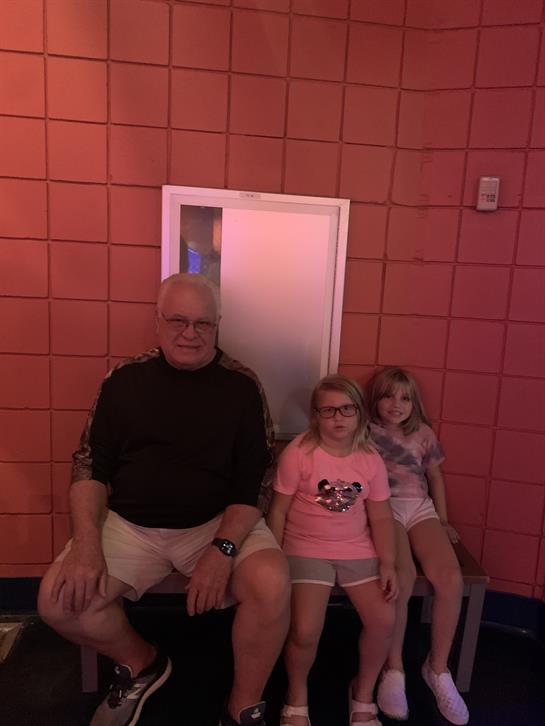 Two young girls are smiling beside their grandfather in a vibrant indoor setting filled with fun.