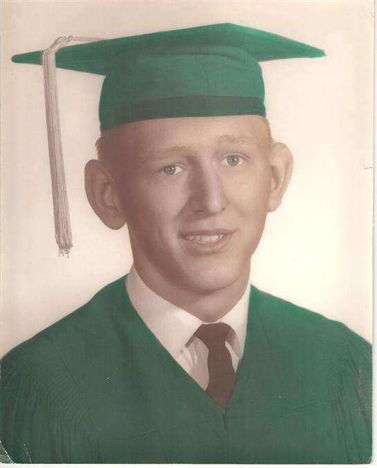 A young man in a green graduation cap and gown smiles confidently at the camera in a studio.
