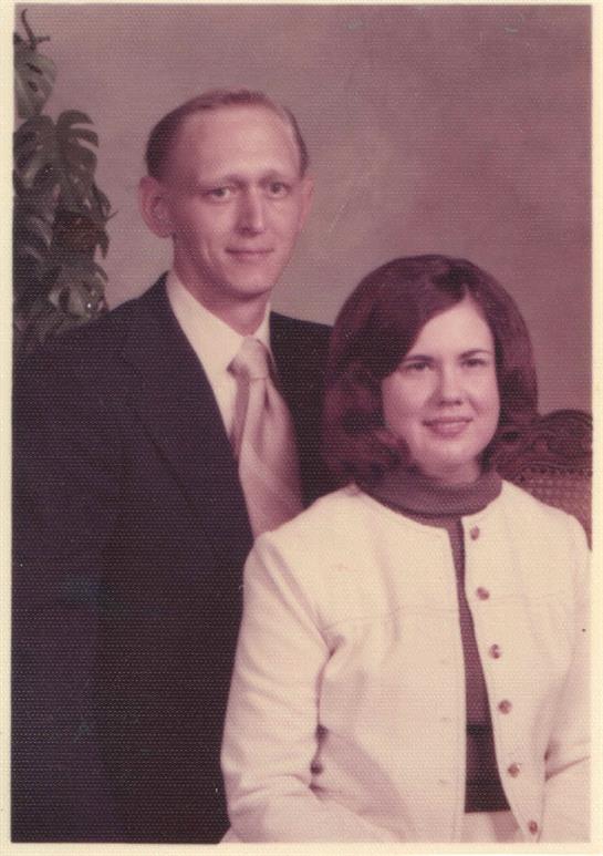 A couple stands closely together, dressed in formal clothes typical of the 1970s era, smiling.
