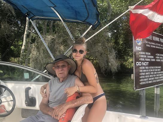 A young woman embraces an elderly man while relaxing on a boat in clear water surrounded by trees.