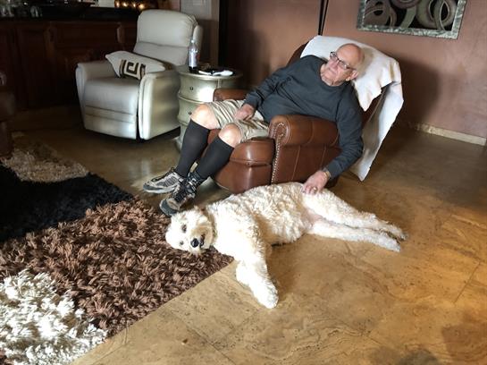 Elderly man rests comfortably in a chair while his dog lounges on a rug nearby.