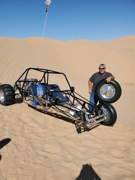 A man stands beside a custom dune buggy in a sunlit desert, showcasing its design.