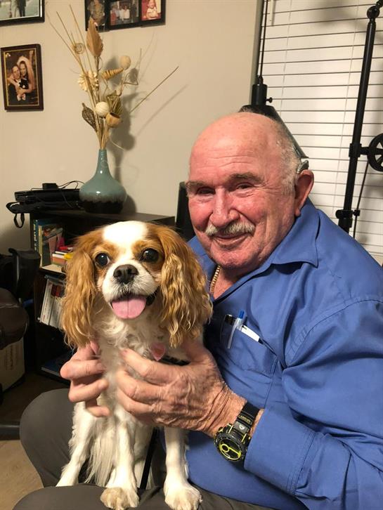 An older man smiles, holding a small dog in a cozy, personalized living space.