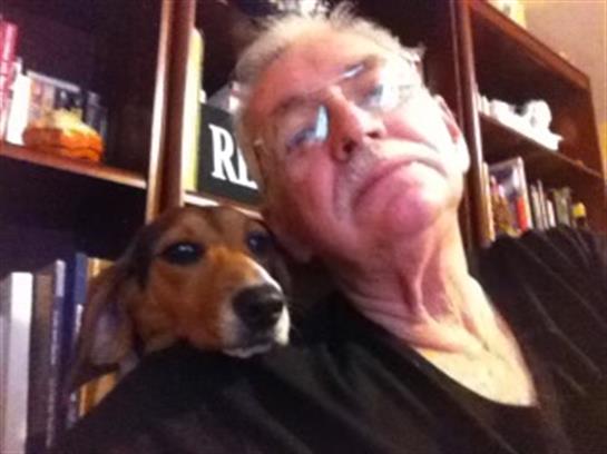 An elderly man relaxes in a library, sharing a warm moment with his dachshund on his shoulder.