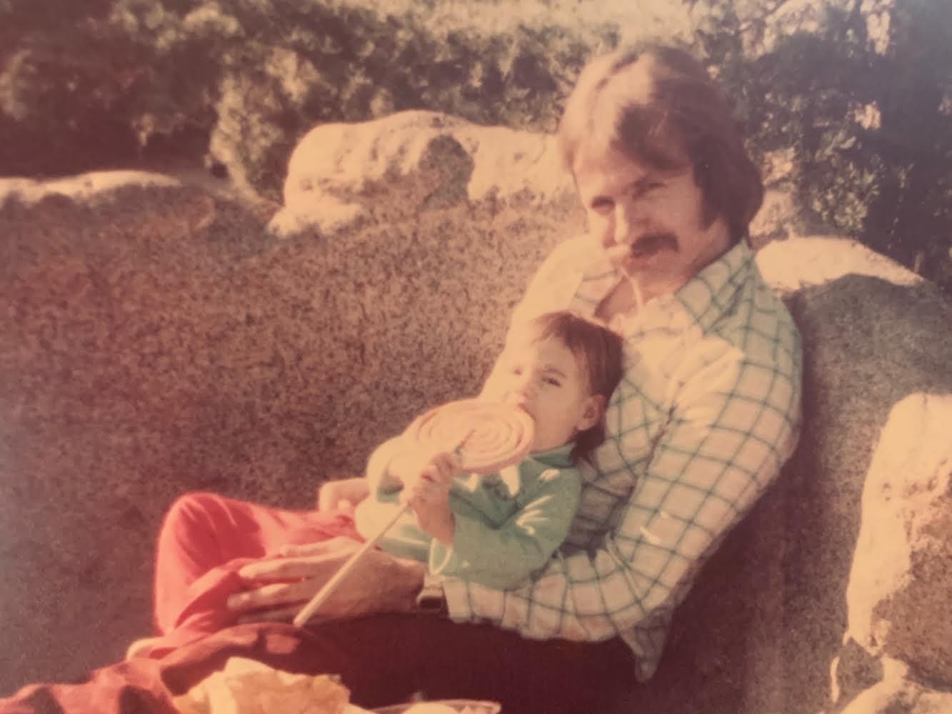 A father embraces his child while enjoying a treat outdoors on a sunny day in the 1970s.