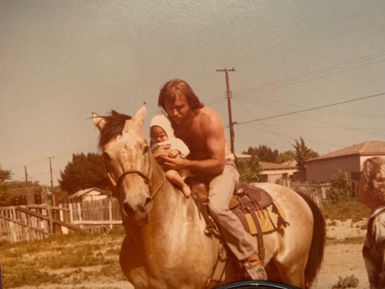 A man holds a baby while riding a horse in a warm suburban area, enjoying a sunny day.