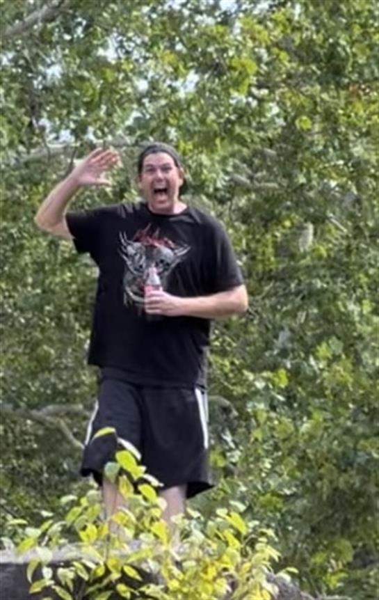 Person stands amidst trees, waving cheerfully and holding a beverage in hand under natural sunlight.