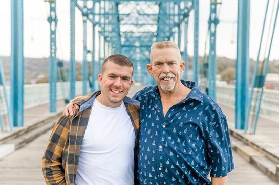 Two men stand side by side on a blue bridge, sharing a joyful moment during fall.