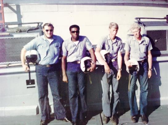 Four pilots in uniforms stand together, holding helmets, near an aircraft at an airfield.